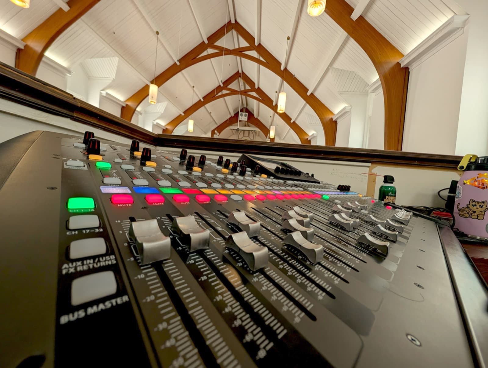 A digital mixing console at the back of a church sanctuary, timber roof arches rising behind it