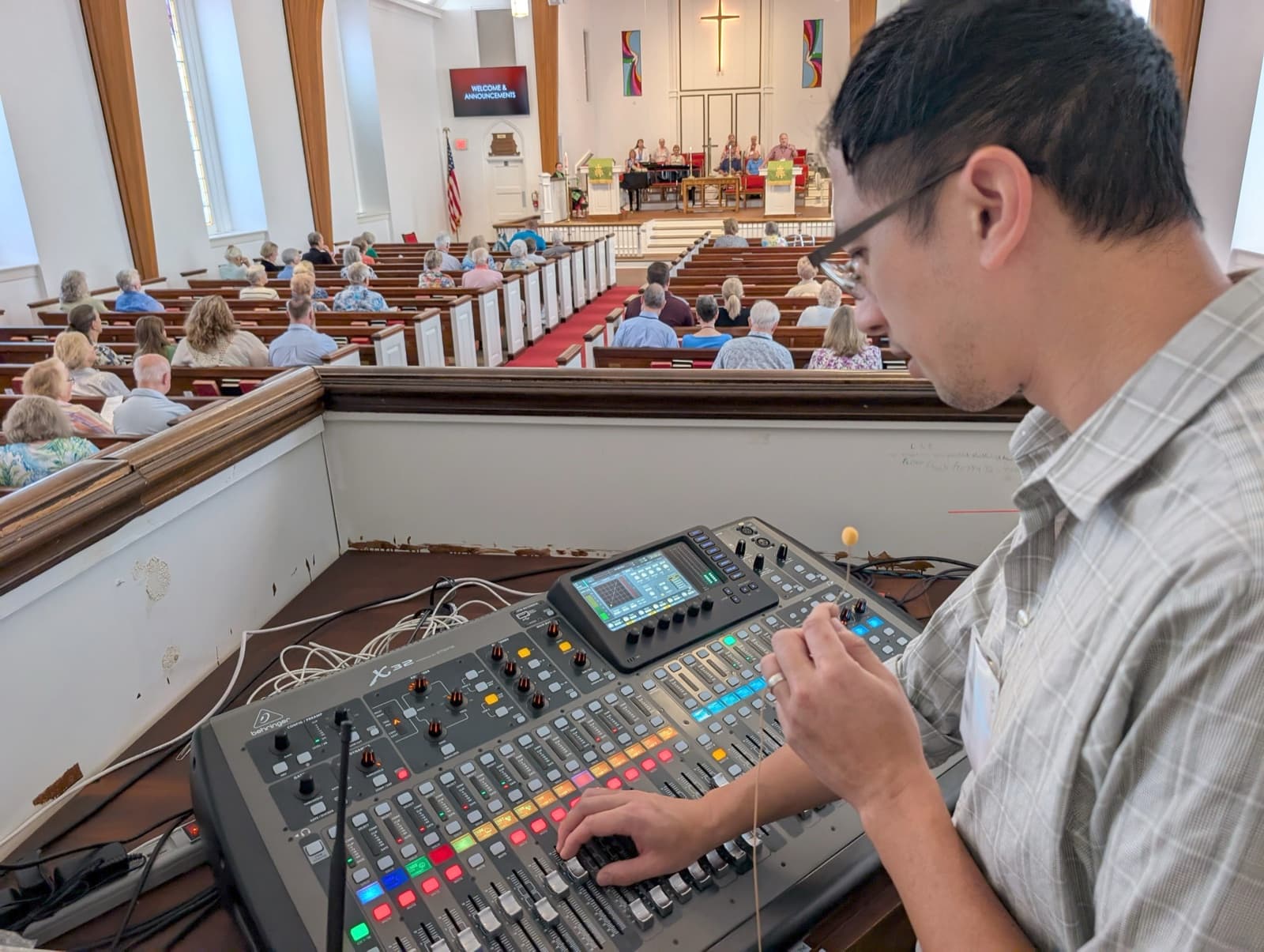Kyle Ng mixing a Sunday service from the balcony of a church sanctuary, hands on a digital console with the congregation seated below
