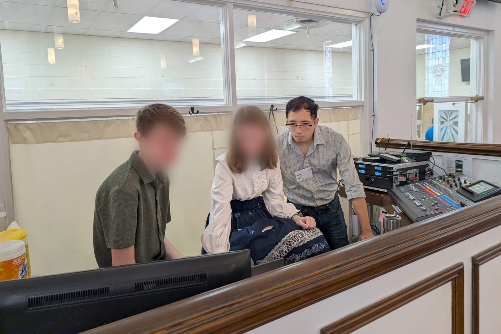 Two church volunteers at a digital console in a sound booth with Kyle Ng standing over their shoulders